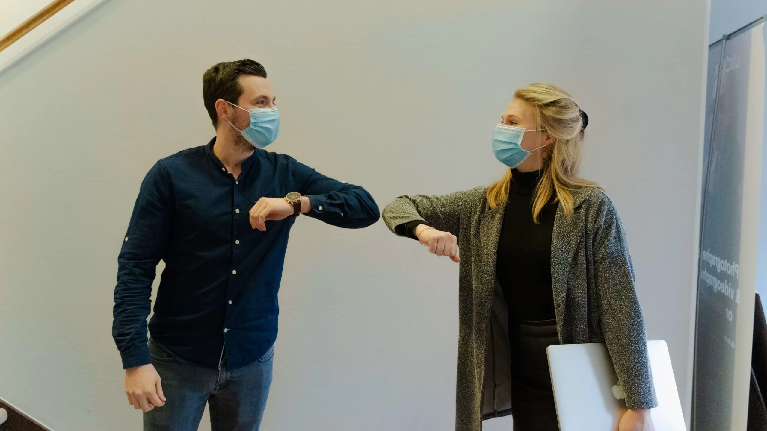 Dos compañeros con mascarilla saludándose con el codo en la oficina, reflejando el uso de apps corporativas en el entorno de trabajo
